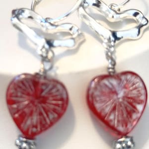 May include: A pair of silver-toned hoop earrings featuring a dangling heart-shaped charm. Each charm is made of translucent red glass with a white radial pattern and is accented with a small, textured silver-toned bead at the bottom.