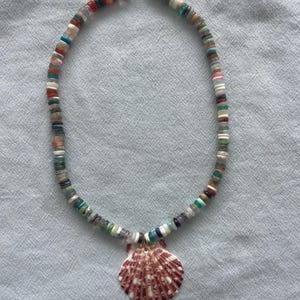 May include: A colorful beaded necklace with a seashell pendant. The necklace features small, flat, multi-colored beads and a large, reddish-brown seashell pendant with white stripes. The necklace is on a light blue background.