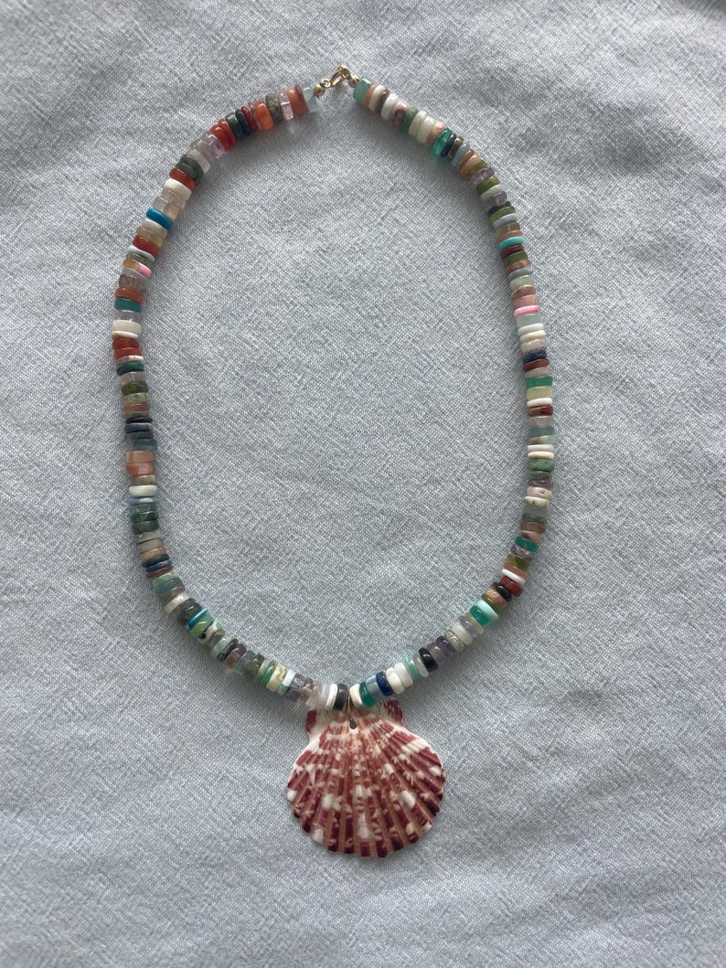 May include: A colorful beaded necklace with a seashell pendant. The necklace features small, flat, multi-colored beads and a large, reddish-brown seashell pendant with white stripes. The necklace is on a light blue background.