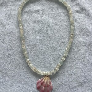 May include: A delicate necklace featuring small, pale green-blue gemstone beads. A pink and white seashell pendant hangs from the center. The necklace is displayed on a light blue fabric background.