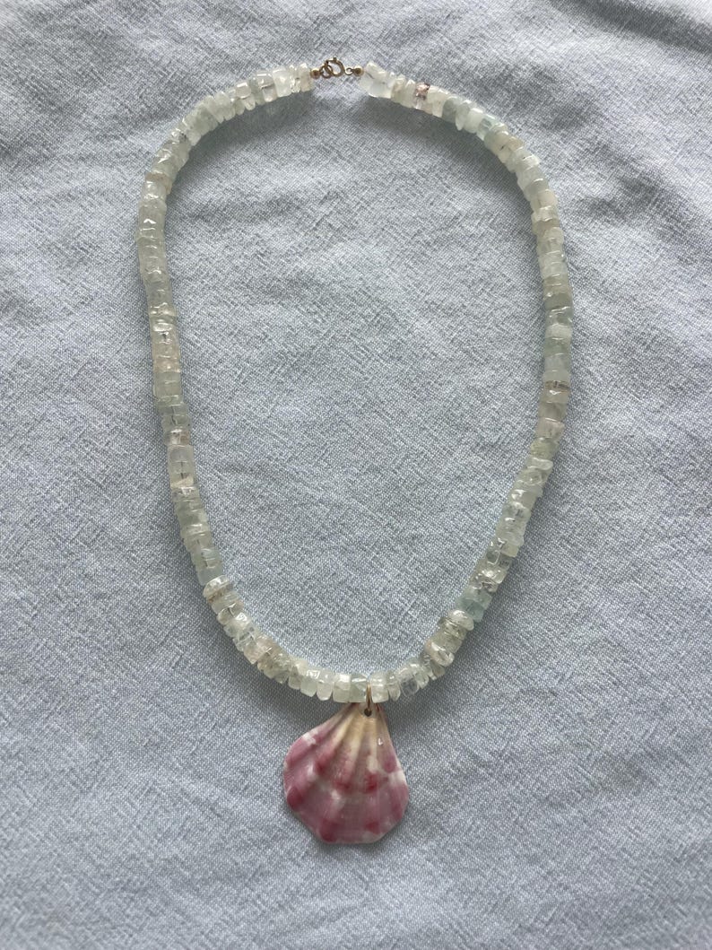 May include: A delicate necklace featuring small, pale green-blue gemstone beads. A pink and white seashell pendant hangs from the center. The necklace is displayed on a light blue fabric background.