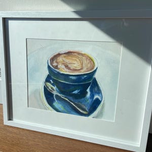 Pu&ograve; includere: Un dipinto ad acquerello incorniciato di una tazza di caff&egrave; blu con una bevanda al caff&egrave; marrone all'interno. La tazza &egrave; appoggiata su un piattino blu con un cucchiaio d'argento.