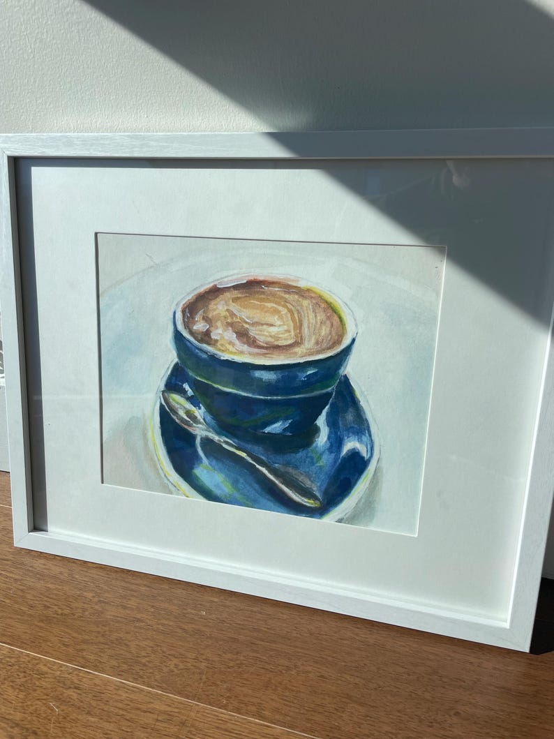 Pu&ograve; includere: Un dipinto ad acquerello incorniciato di una tazza di caff&egrave; blu con una bevanda al caff&egrave; marrone all'interno. La tazza &egrave; appoggiata su un piattino blu con un cucchiaio d'argento.