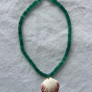 May include: A green beaded necklace with a seashell pendant. The necklace features small, round green beads and a white seashell with reddish-brown markings. The necklace is displayed on a light gray fabric.