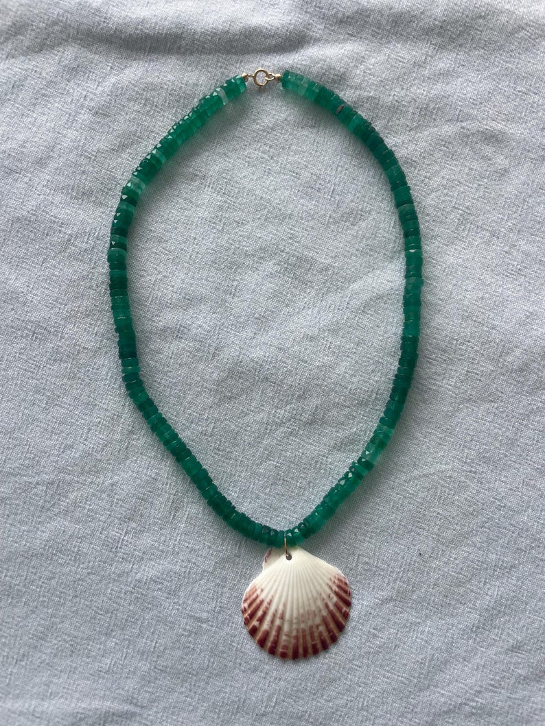 May include: A green beaded necklace with a seashell pendant. The necklace features small, round green beads and a white seashell with reddish-brown markings. The necklace is displayed on a light gray fabric.