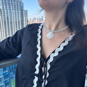 May include: A black top with white trim and a seashell necklace. The necklace features a white seashell pendant and a beaded strand. The top has a V-neck with a tie closure.