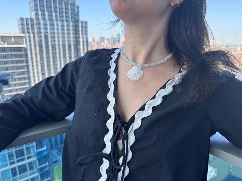 May include: A black top with white trim and a seashell necklace. The necklace features a white seashell pendant and a beaded strand. The top has a V-neck with a tie closure.