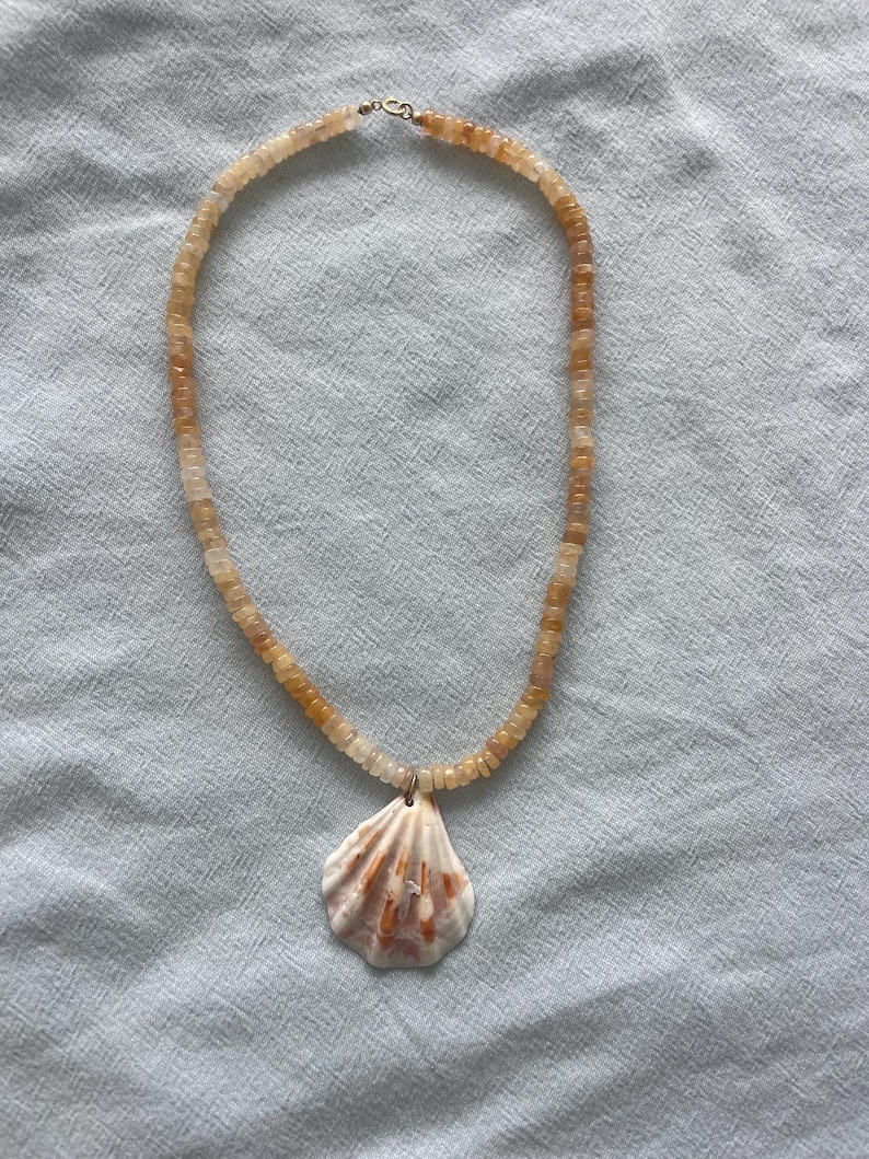 May include: A necklace featuring a shell pendant and a beaded strand. The shell pendant is white with orange and brown markings. The beaded strand is made of small, square, amber-colored beads. The necklace is displayed on a light blue surface.