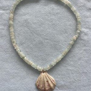 May include: A necklace featuring a strand of light green, square-cut beads. A natural seashell pendant with pink and beige hues hangs from the center. The necklace is secured with a gold-colored clasp.