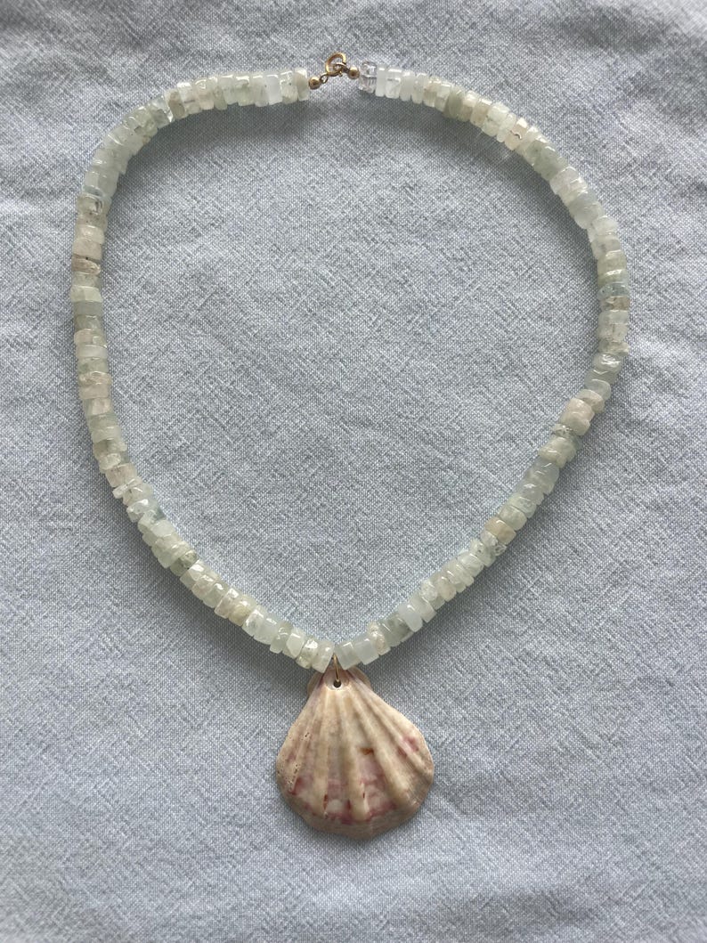 May include: A necklace featuring a strand of light green, square-cut beads. A natural seashell pendant with pink and beige hues hangs from the center. The necklace is secured with a gold-colored clasp.