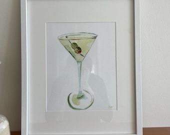 May include: A framed watercolor painting of a martini glass with a green olive. The glass is filled with a light green liquid and the olive is resting on the rim of the glass. The painting is on a white background and is framed in a white frame.