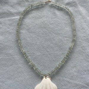 May include: A delicate necklace featuring a strand of light green, small, square-cut beads. A white seashell pendant hangs from the necklace. The necklace is secured with a gold-colored clasp. The necklace is displayed on a light blue fabric.