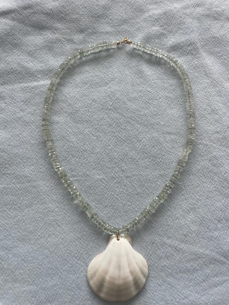 May include: A delicate necklace featuring a strand of light green, small, square-cut beads. A white seashell pendant hangs from the necklace. The necklace is secured with a gold-colored clasp. The necklace is displayed on a light blue fabric.