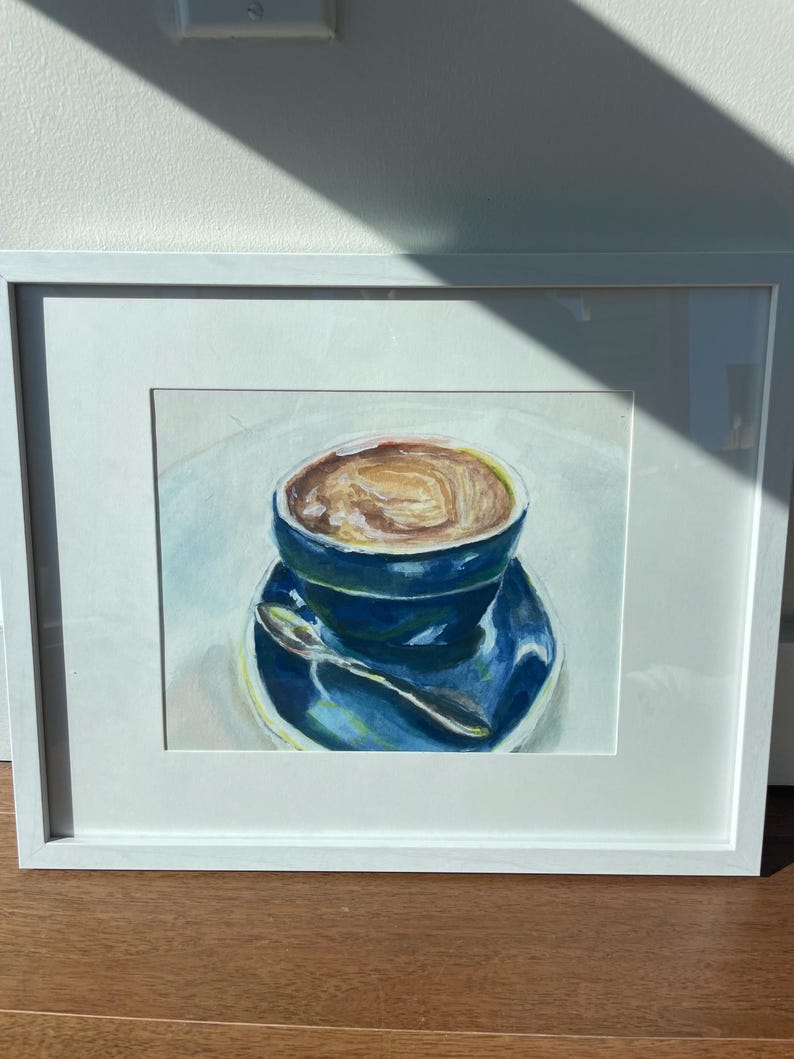 Pu&ograve; includere: Un dipinto incorniciato di una tazza di caff&egrave; blu con una bevanda al caff&egrave; marrone all'interno. La tazza &egrave; su un piattino blu con un cucchiaio d'argento.