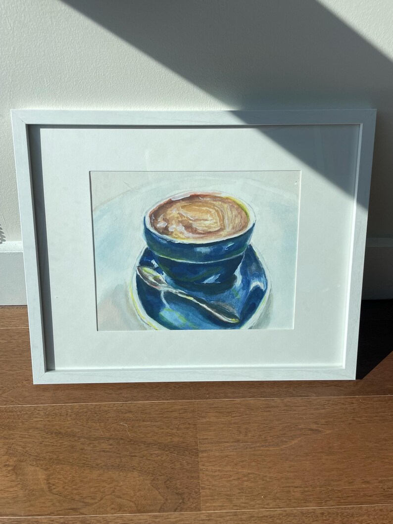 Pu&ograve; includere: Un dipinto incorniciato di una tazza di caff&egrave; blu con un liquido marrone al suo interno. La tazza &egrave; su un piattino bianco con un cucchiaio d'argento.