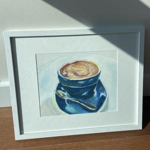 Pu&ograve; includere: Un dipinto ad acquerello incorniciato di una tazza di caff&egrave; blu con una bevanda al caff&egrave; marrone all'interno. La tazza &egrave; su un piattino blu con un cucchiaio d'argento.