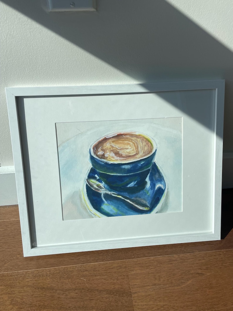 Pu&ograve; includere: Un dipinto ad acquerello incorniciato di una tazza di caff&egrave; blu con una bevanda al caff&egrave; marrone all'interno. La tazza &egrave; su un piattino blu con un cucchiaio d'argento.