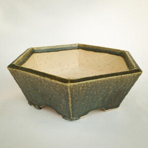 May include: A hexagonal ceramic planter with a textured, dark olive-green exterior glaze and a smooth, speckled cream-coloured interior. The vessel features a wide, open top rim and is supported by small, integrated feet at the base.