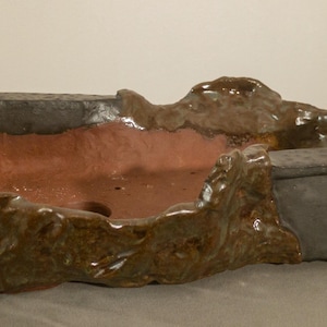 May include: A rectangular bonsai pot with a textured, dark grey rim and a reddish-brown interior. The pot has a unique, organic shape with raised, textured areas in shades of brown and green. The pot is approximately 30 cm long.