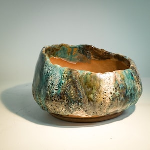 May include: A round ceramic bowl with an irregular, textured rim and a smooth, light brown interior. The exterior features a mottled glaze in shades of teal, blue, green, and brown, with a porous, cratered surface texture throughout the outer body.