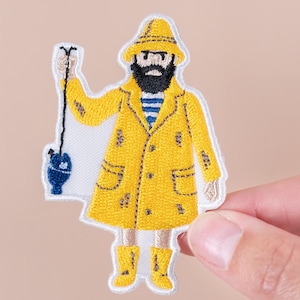 May include: Embroidered patch featuring a cartoon fisherman in a yellow raincoat and hat, holding a fishing rod with a blue and white buoy. The patch has a white border and is held by a hand against a neutral background.