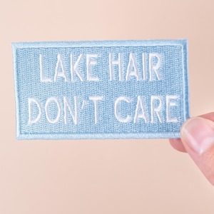 May include: A light blue embroidered patch with the text "LAKE HAIR DON'T CARE" in white. The patch is rectangular with a light blue border and is held by a hand against a peach background.