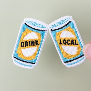 May include: Two embroidered patches in the shape of beer cans, one reading "DRINK" and the other "LOCAL". The patches are blue, white, yellow, and black, held by a hand against a light green background. The patches are likely for clothing or accessories.