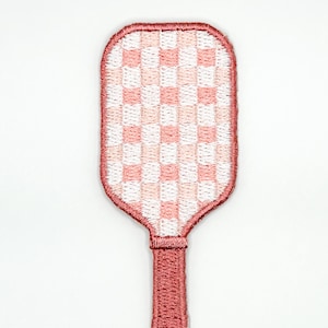 May include: An embroidered patch shaped like a pickleball paddle featuring a white and light pink checkered pattern on the head. The paddle head is outlined with a thick pink border, and the handle is solid pink with a matching embroidered edge.