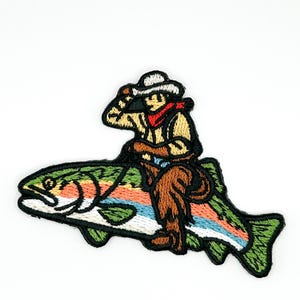 May include: An embroidered patch featuring a figure wearing a white cowboy hat, tan shirt, red neckerchief, and brown pants, riding on the back of a large rainbow trout. The fish has green scales, a white belly, and a pinkish-orange lateral stripe.