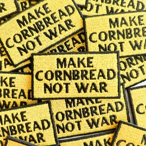 May include: A pile of yellow fabric patches with black embroidered text that reads "Make Cornbread Not War".