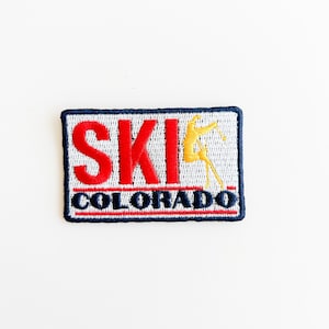 Op de afbeelding: Een witte geborduurde patch met blauwe en rode letters die "SKI COLORADO" lezen. In de rechterbovenhoek van de patch staat een geel silhouet van een skiër.