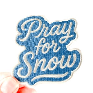 May include: A blue patch with white embroidered text that reads "Pray for Snow." The patch has a denim-like texture and a white border. The patch is held up against a white background.
