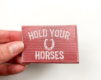 Pode incluir: Um patch retangular rosa empoeirado com texto bordado branco que diz "HOLD YOUR HORSES" com um símbolo de ferradura. O patch é mantido contra um fundo branco.