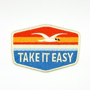 May include: A hexagonal embroidered patch featuring a sunset landscape with a white seagull flying above horizontal stripes of red, orange, and cream, over a blue base with the white text TAKE IT EASY stitched in a bold, sans-serif font.
