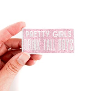 May include: Pink fabric patch with white embroidered text that reads "Pretty Girls Drink Tall Boys".
