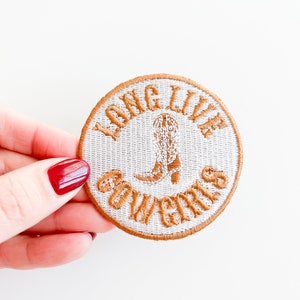 Puede incluir: Un parche redondo bordado con las palabras "LONG LIVE COWGIRLS" en un diseño circular. El parche presenta una bota de vaquero marrón en el centro y un borde marrón. El parche está sostenido por una mano con esmalte de uñas rojo.