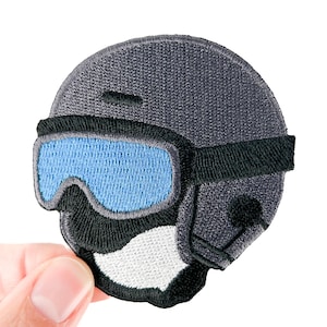 May include: Embroidered patch featuring a gray ski helmet with black goggles and a white mouth guard. The goggles have blue lenses. The patch is round and has a textured appearance.
