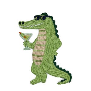 Puede incluir: Un parche bordado que muestra un caimán verde con gafas de sol negras. El caimán está de pie y sostiene una copa de martini con una aceituna en la mano. El diseño presenta un vientre de color tostado claro y un dorso verde oscuro.