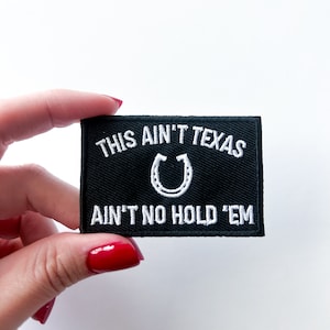 Puede incluir: Parche bordado negro con texto blanco que dice "This ain't Texas ain't no hold 'em" con una herradura en el centro.