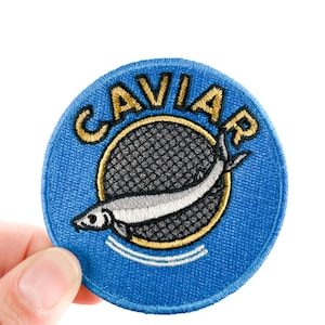 Pode incluir: Um patch redondo bordado em azul com a palavra "CAVIAR" em letras douradas. O patch apresenta um peixe estilizado dentro de um círculo com borda dourada e um padrão de rede cinza. O patch é segurado por uma mão em um fundo branco.
