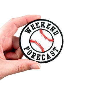以下が含まれることがあります： 縁が黒い円形の白い刺繍パッチ。円には「Weekend Forecast」という文字が黒で書かれており、中央には赤い野球ボールがあります。