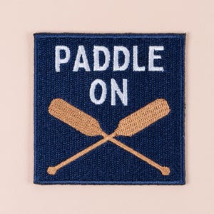 May include: A navy blue patch with the words "PADDLE ON" in white block letters above two crossed wooden paddles. The paddles are light brown and have a textured appearance.