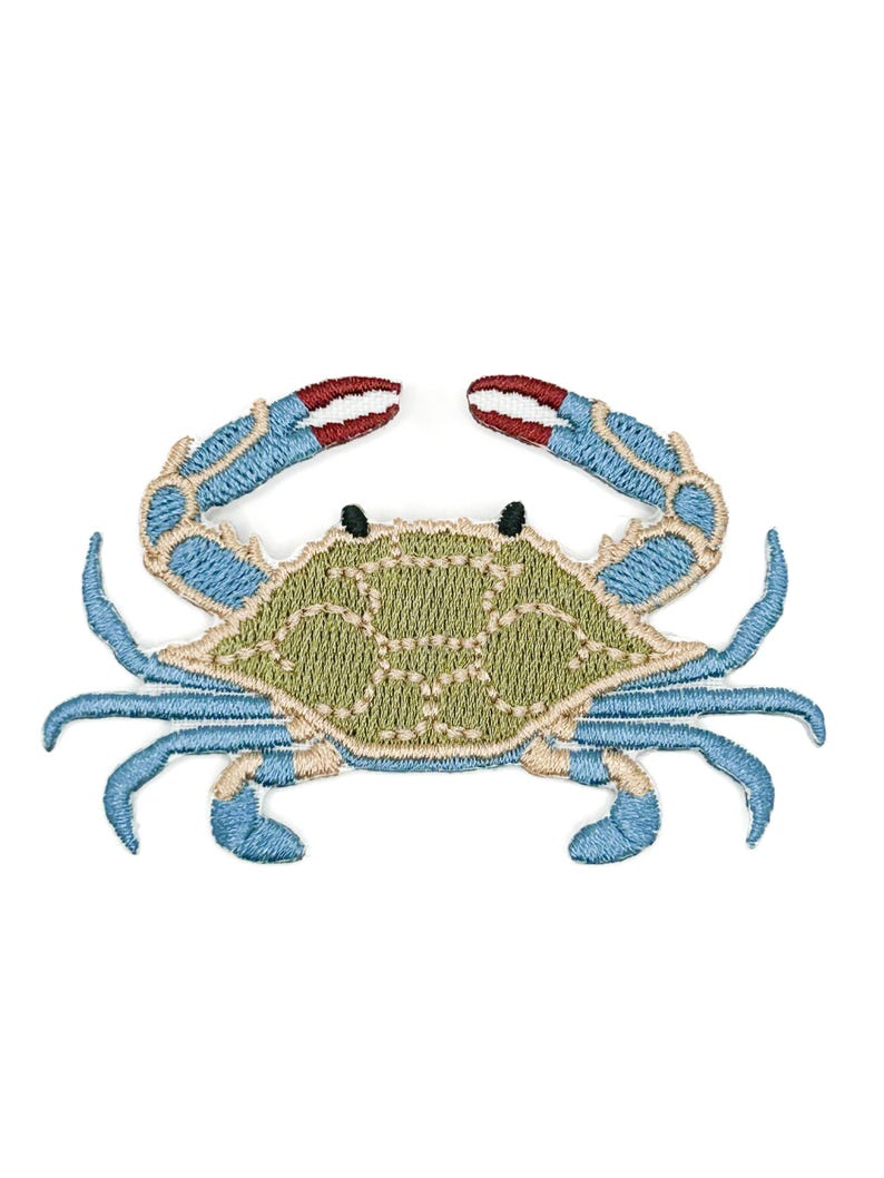 May include: An embroidered iron-on patch shaped like a blue crab. The crab features a textured olive green shell with light tan outlines, bright blue legs and claws, and small black eyes. The tips of the claws are accented with white and red thread.