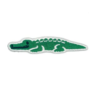Peut inclure: Un écusson brodé en forme d'alligator vert avec une bouche et un œil vert foncé, bordé d'une couture blanche. Le motif représente l'alligator de profil, tourné vers la gauche, avec un corps texturé et une longue queue pointue.