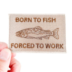 Może przedstawiać: Prostokątna naszywka w kolorze beżowym z brązowym haftowanym tekstem i wzorem ryby. Tekst brzmi "BORN TO FISH" nad rybą i "FORCED TO WORK" poniżej. Naszywka jest trzymana przez dłoń na białym tle.