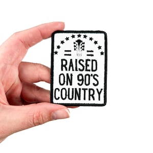 Może przedstawiać: Biała naszywka do prasowania z czarnym haftem, na której widnieje napis "Raised on 90's Country" z rzędem gwiazd nad i pod tekstem.