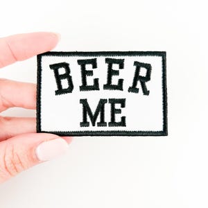 Puede incluir: Parche blanco para planchar con texto bordado en negro que dice "BEER ME".