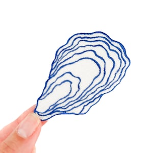 Può includere: Una toppa di tessuto bianco con un disegno a conchiglia blu. La toppa presenta un contorno ricamato dettagliato dell'interno della conchiglia, con linee concentriche che creano un effetto strutturato. La toppa è tenuta su uno sfondo bianco.