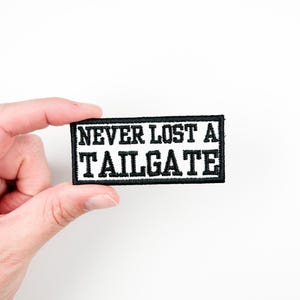 May include: A rectangular black and white patch with the text "NEVER LOST A TAILGATE" embroidered in white block letters. The patch is held against a white background.