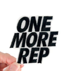 May include: A white embroidered patch with the words "ONE MORE REP" in bold, black letters. The patch has a rectangular shape with rounded corners. The text is stitched with a thick, textured thread.